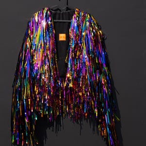 May include: A jacket covered in long, reflective, multi-colored metallic fringe in shades of blue, purple, green, gold, and red. The garment features a black interior lining and a small orange label with the text House of Mairzel at the neck.