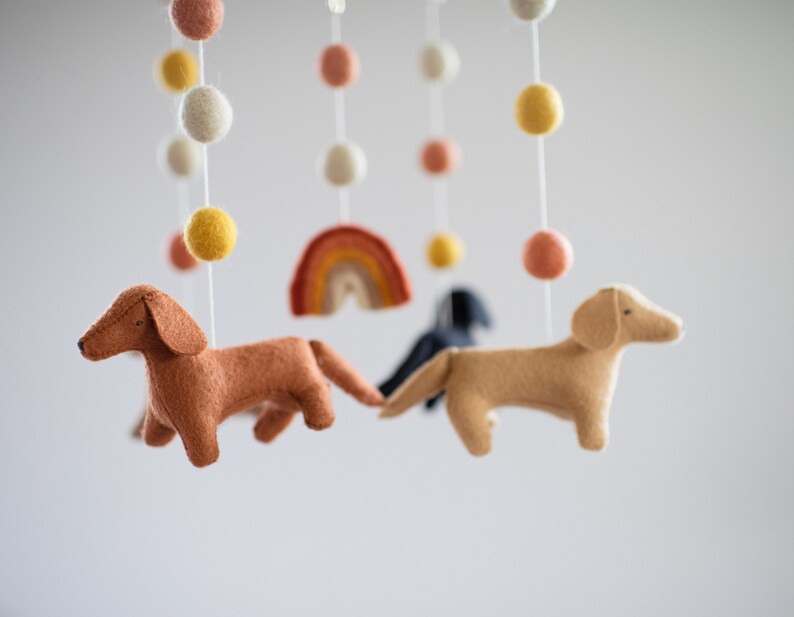 May include: A felt baby mobile with a rainbow, white, yellow, and pink pom poms, and three felt dachshund dogs in brown, tan, and black.