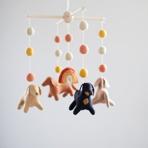 May include: A baby mobile with felt dog and rainbow decorations hanging from a wooden frame. The mobile features colorful pom poms in shades of yellow, orange, and white.