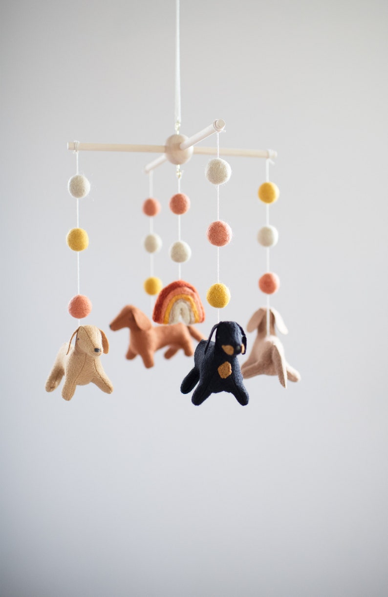 May include: A baby mobile with felt dog and rainbow decorations hanging from a wooden frame. The mobile features colorful pom poms in shades of yellow, orange, and white.