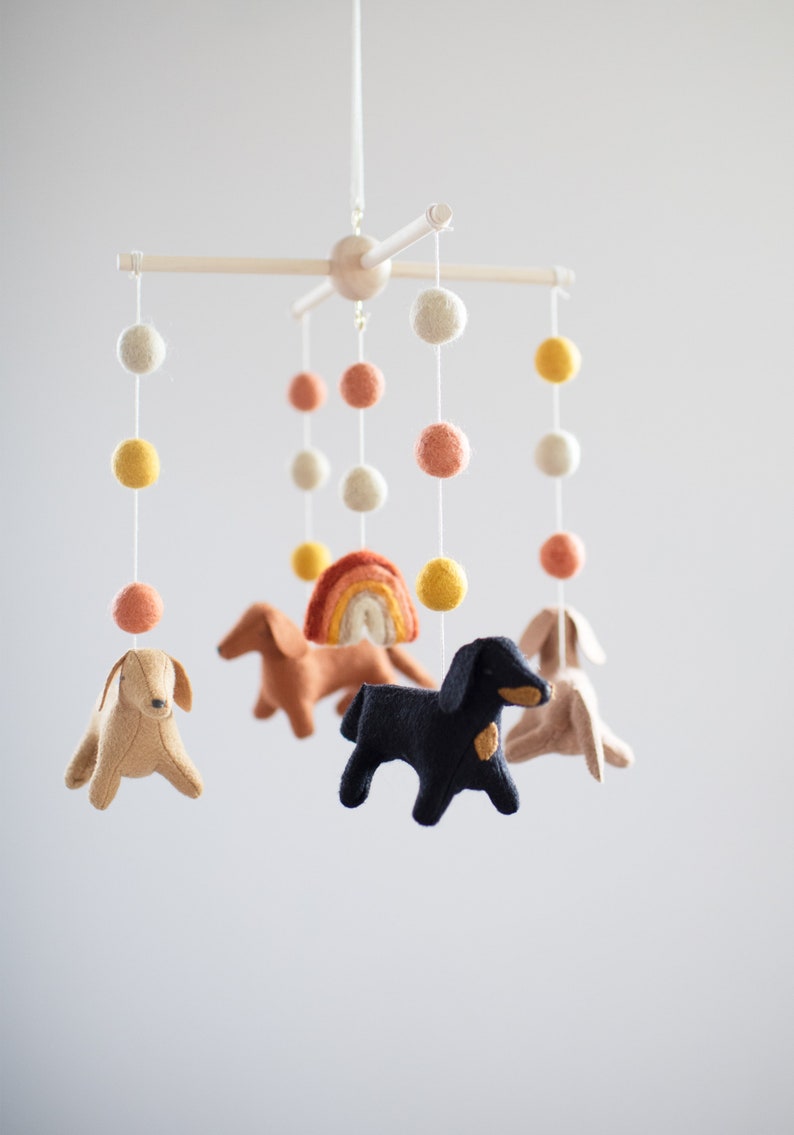 May include: A felt baby mobile with a rainbow, three brown and black dachshund dogs, and a white, yellow, and orange pom poms.