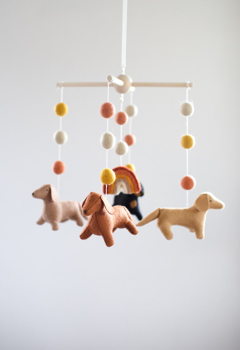 May include: A wooden mobile with felt dachshund dogs, a rainbow, and colorful pom poms hanging from it.