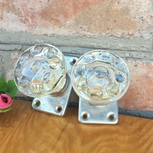 May include: Two clear glass door knobs with a textured surface, mounted on silver metal plates. The knobs are on a wooden surface.