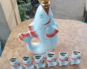 May include: A white ceramic fish-shaped decanter with red painted scales and fins, topped with a gold-colored spherical stopper, accompanied by six matching small fish-shaped ceramic cups, each featuring red painted details and a hollow center.
