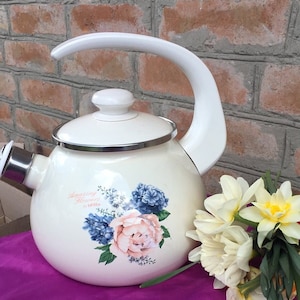 May include: White enamel teapot with a floral design featuring pink, blue, and green flowers. The teapot has a silver trim and the text "Amazing Flowers by Lilla" on the side. The teapot is sitting on a purple tablecloth.