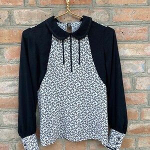 Könnte beinhalten: Eine weiße Bluse mit schwarzem Muster, schwarzem Kragen und langen Ärmeln. Die Ärmel haben einen schwarzen Besatz und Knöpfe an den Manschetten.