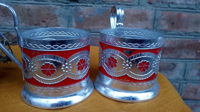 May include: Two silver-colored metal tea glass holders with red enamel and floral designs. The holders have a handle on the side.