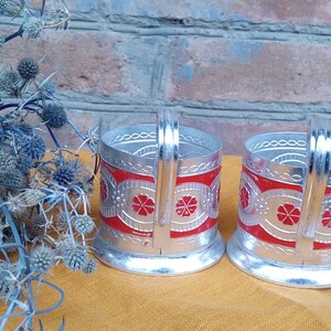 May include: Two silver-colored metal teacup holders with red and silver floral designs. The holders have a handle on the side and a rounded base.
