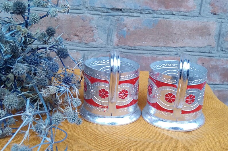 May include: Two silver-colored metal teacup holders with red and silver floral designs. The holders have a handle on the side and a rounded base.