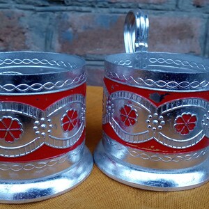 May include: Two silver-colored metal tea glass holders with red enamel and floral designs. The holders have a handle on the side.