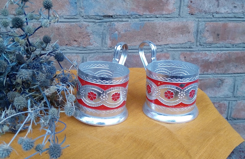 May include: Two silver metal cup holders with red and silver floral designs. The holders have a handle and are sitting on a yellow surface.