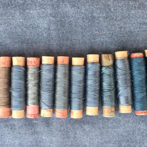 Könnte beinhalten: Eine Sammlung von 15 Garnrollen in verschiedenen Blautönen, Grüntönen und Brauntönen. Die Spulen sind aus Holz und haben ein Papieretikett.