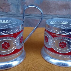 May include: Two silver-colored metal tea glass holders with red and silver floral designs. The holders have a handle and a round base.