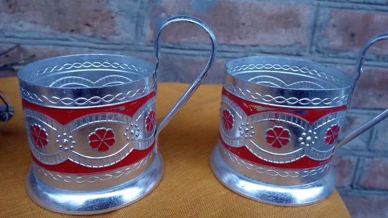 May include: Two silver-colored metal tea glass holders with red and silver floral designs. The holders have a handle and a round base.