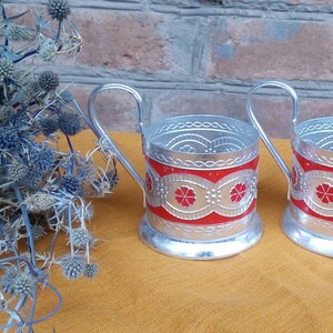 May include: Two silver metal cup holders with red and white floral designs. The holders have a handle and are sitting on a yellow surface.