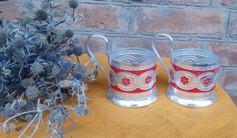 May include: Two silver metal cup holders with red and white floral designs. The holders have a handle and are sitting on a yellow surface.