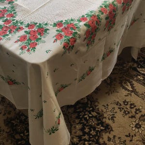 May include: White rectangular tablecloth with a floral pattern of red roses and green leaves. The roses are clustered in a border around the edges of the tablecloth. The tablecloth is draped over a wooden table.