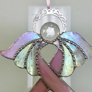 May include: A stained glass angel ornament with a pink ribbon, iridescent wings, and a silver halo. The angel has a faceted crystal head and is suspended by a pink ribbon. The wings have a pastel color scheme.