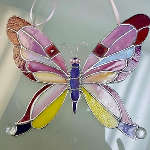 May include: A stained glass butterfly with pink, yellow, red, and purple glass pieces. The butterfly has clear crystal accents and a pink ribbon for hanging.