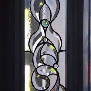 May include: A stained glass window panel with a decorative design of overlapping teardrop shapes. The glass is clear with a subtle iridescent sheen and features small, clear glass jewels.