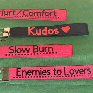 May include: Four red and black fabric keychains with different phrases printed on them. The phrases are "Hurt/Comfort", "Kudos", "Slow Burn", and "Enemies to Lovers".