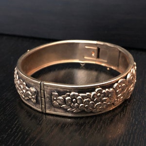 May include: A gold-toned hinged bangle bracelet with a floral design. The bracelet is made of metal and has a smooth finish.