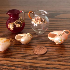 May include: Set of five miniature glass and shell pitchers. The pitchers are decorated with seashells and flowers. The pitchers are in various colors, including red, clear, and pink.