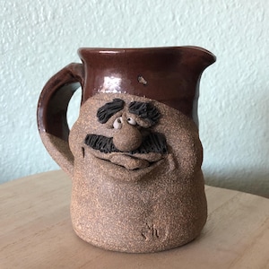 May include: A brown and tan ceramic pitcher with a whimsical face. The pitcher has a brown glaze on the top half and a tan glaze on the bottom half. The face has a mustache, eyebrows, and two eyes.