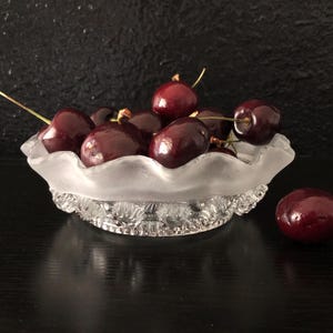 May include: A clear, frosted glass bowl filled with ripe, dark red cherries. The bowl has a scalloped rim and a decorative, textured base. One cherry sits outside the bowl on a dark surface.
