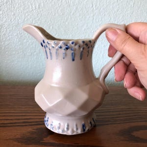 May include: A small, white ceramic creamer with a blue and white floral design. The creamer has a handle and a rounded body.