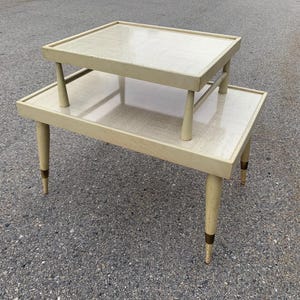 May include: A two-tiered, vintage side table with a light beige finish. The table has a rectangular top and a smaller, elevated top. The legs are tapered and have brass accents. A classic piece of mid-century modern furniture.