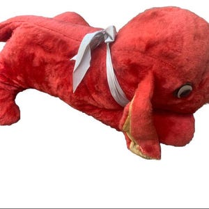 May include: A red plush toy dog with a white ribbon around its neck. The dog is lying on its side with its head turned to the side.