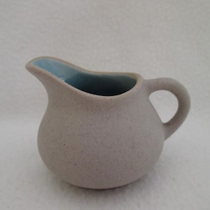 May include: A small, speckled, beige ceramic creamer with a light blue interior. The creamer has a rounded body, a curved spout, and a small handle. The design is simple and classic, suitable for serving cream or milk.