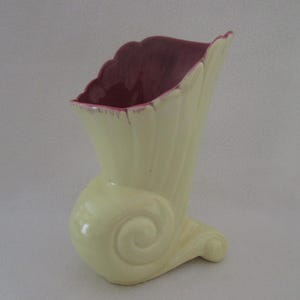 May include: A yellow ceramic vase with a pink interior and a unique, stylized design. The vase has a curved base and a tall, narrow opening.
