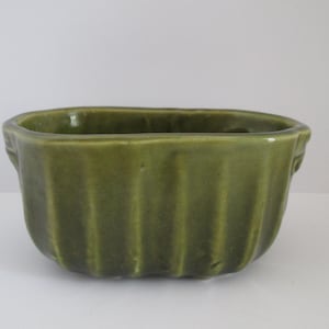 May include: A small, olive green ceramic planter with a rectangular shape and vertical ribbed detailing. The planter has a glossy finish and small handles on each side. Ideal for small plants or succulents.