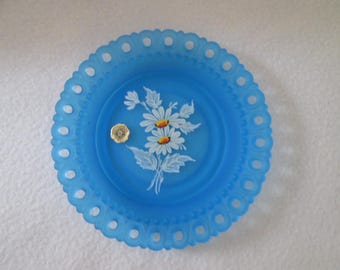 Può includere: Un piatto di vetro blu con un motivo di pizzo bianco attorno al bordo. Il piatto presenta un disegno dipinto a mano di margherite bianche con centri gialli.