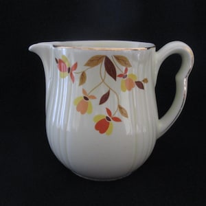 May include: A white ceramic creamer with a gold rim and a floral pattern of orange, yellow, and brown leaves. The creamer has a rounded body and a handle.