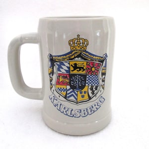 May include: A light gray ceramic beer mug with a sturdy handle. The front features a colorful heraldic crest with a crown on top and the text KARLSBERG printed in blue capital letters below the shield, which contains various geometric patterns.