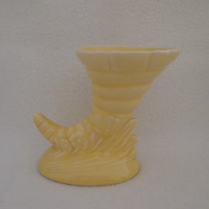 May include: A yellow ceramic vase shaped like a cornucopia. The vase has a smooth, glossy finish and a slightly textured surface.