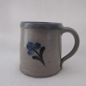May include: A stoneware mug with a gray body and a blue rim and handle. A blue floral design with a green stem is painted on the front of the mug. The mug has a cylindrical shape and a curved handle.