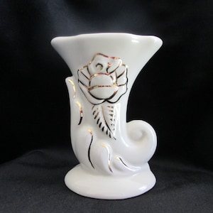 May include: White ceramic vase with a gold rose design. The vase is shaped like a horn and has a wide opening at the top.