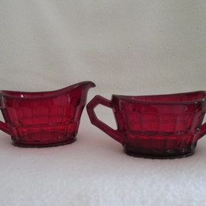 May include: A ruby red glass creamer and sugar bowl set. The creamer has a spout and a handle, while the sugar bowl has two handles. Both pieces have a textured, geometric pattern. The set is likely vintage.