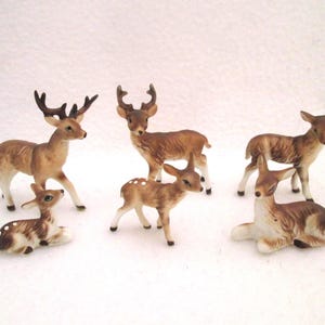 May include: A collection of six ceramic deer figurines in various poses. The set includes two standing bucks with antlers, two standing fawns with white spots, and two fawns resting in a lying position. Each figurine features brown and white coloring.