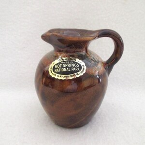 May include: A small brown glazed ceramic pitcher with a rounded body and a single handle. An oval sticker on the front reads: Dryden Made in Hot Springs National Park From Ark. Clay and Crystal Quartz. The finish is glossy with dark brown swirls.