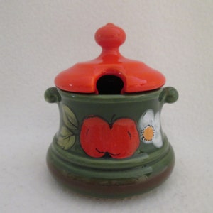 May include: A green ceramic sugar bowl with a red lid and a floral design. The lid has a small hole for pouring.