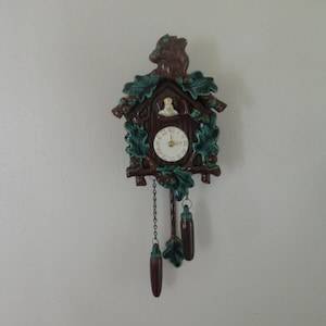 May include: A brown and green cuckoo clock with a squirrel on top and a bird on the face. The clock has a chain with two brown acorn-shaped weights.