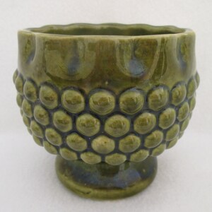 May include: A green ceramic planter with a textured, bubble-like design. The planter has a wide, round base and a flared rim.