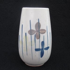 May include: A white ceramic vase with a rounded shape, decorated with blue vertical lines and two stylized floral designs in brown and blue. The vase has a speckled texture and a red base.