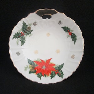 May include: A white ceramic serving dish with a gold rim and a Christmas theme. The dish features a poinsettia flower, holly leaves, and snowflakes. It has a handle on the top.