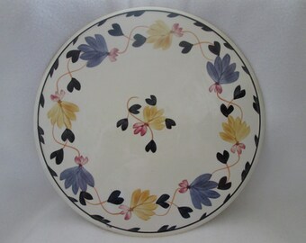 May include: A white ceramic plate with a black, blue, yellow, and pink floral design. The design features a repeating pattern of hearts and leaves around the edge of the plate.